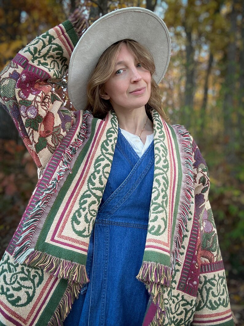 May include: A beige hat and a patterned shawl with green, burgundy, and beige floral and geometric designs. The shawl has a fringed edge. The person is wearing a blue denim dress.