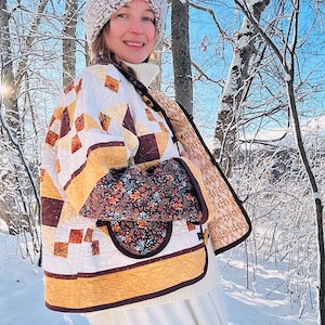 May include: A quilted jacket with a brown, white, and gold patchwork design. The jacket has a brown collar and a floral print lining. The model is wearing a white knit hat and a white sweater.