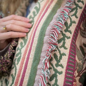 May include: Close-up of a woven textile with a repeating floral pattern in green, burgundy, and beige. The textile has vertical stripes and a fringe of red, white, and green threads. The textile is likely a blanket or throw.