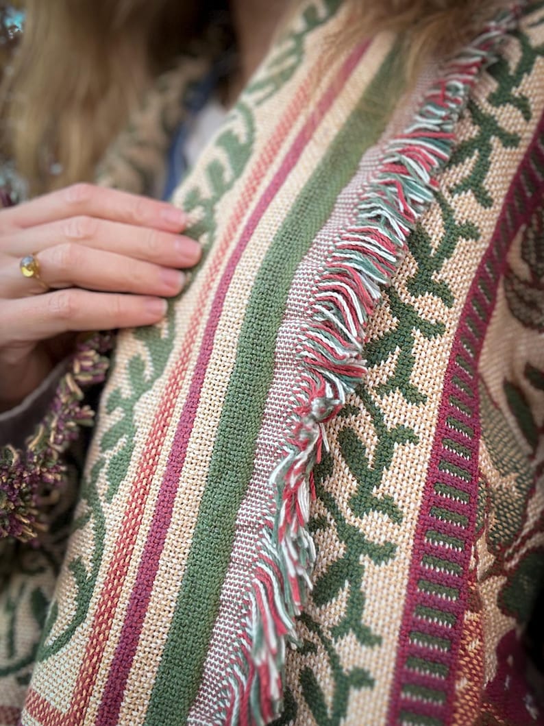 May include: Close-up of a woven textile with a repeating floral pattern in green, burgundy, and beige. The textile has vertical stripes and a fringe of red, white, and green threads. The textile is likely a blanket or throw.
