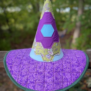 May include: A handmade witch hat with a quilted design. The hat features a wide, circular brim in purple with a floral pattern and a green trim. The cone-shaped crown is adorned with purple and blue geometric shapes and a light green floral fabric.