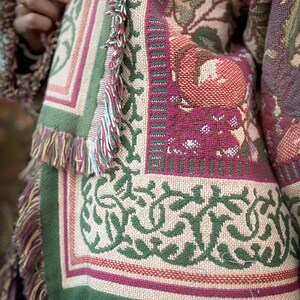 May include: Close-up of a tapestry-style textile with a floral and geometric pattern. The color palette includes beige, green, burgundy, and touches of red. The edges are finished with fringe. The textile appears to be part of a larger garment or decorative item.