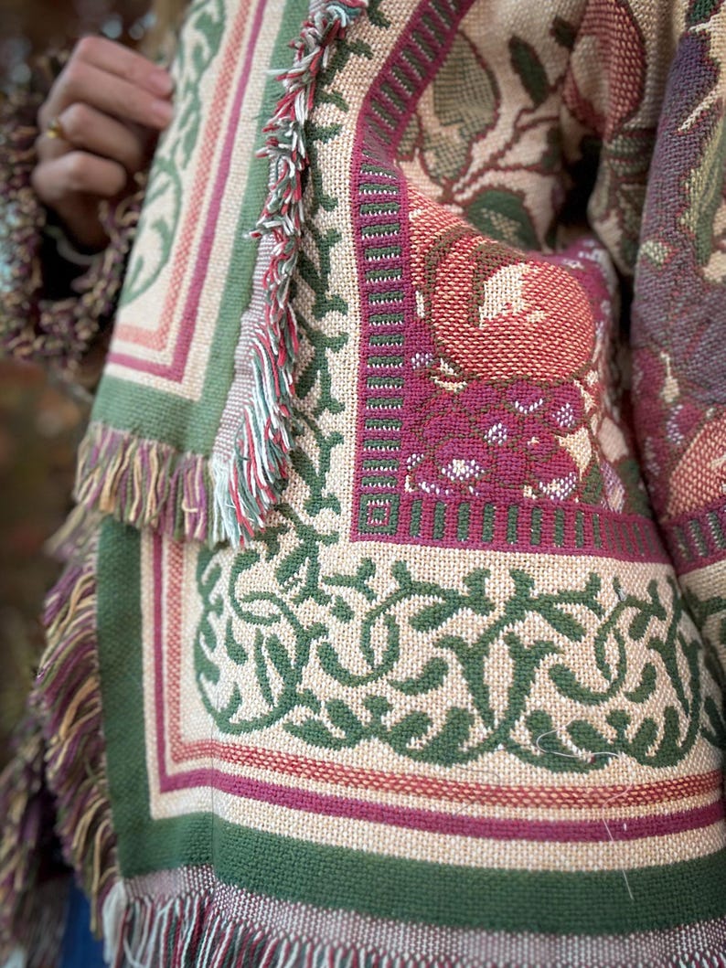 May include: Close-up of a tapestry-style textile with a floral and geometric pattern. The color palette includes beige, green, burgundy, and touches of red. The edges are finished with fringe. The textile appears to be part of a larger garment or decorative item.