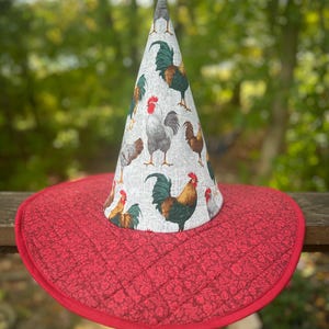 May include: A whimsical, handmade hat with a conical crown and a wide, round brim. The crown features a repeating pattern of colorful roosters on a light background. The brim is a quilted, deep red fabric with a floral design.