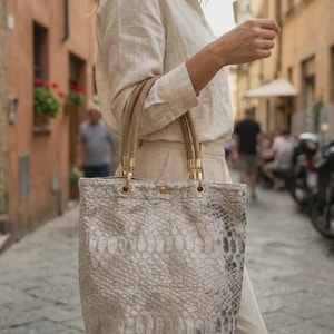 May include: A tote bag featuring a textured snakeskin pattern in shades of beige, grey, and white. The bag has two braided tan handles attached with gold-toned metal hardware and a single gold-toned snap closure at the top centre of the opening.