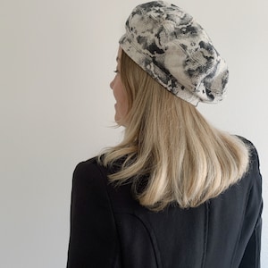 May include: A snake print beret in shades of gray and cream. The beret is worn with a black blazer. The hat has a rounded crown and a brim that sits close to the head. The beret is made of a soft, textured fabric.