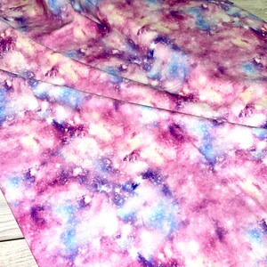 Könnte beinhalten: Lila, blauer und weißer Galaxie-Druckstoff mit weißem Hintergrund. Der Stoff hat eine weiche, gebürstete Textur.