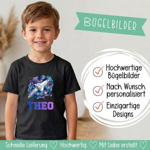 Op de afbeelding: Een zwart T-shirt met korte mouwen en een afbeelding van een spaceshuttle die door een kleurrijke nevel met planeten vliegt. Onder de afbeelding staat de naam THEO in blauwe en paarse blokletters gedrukt op een kind.