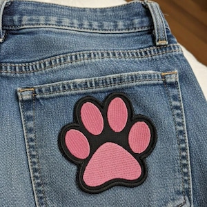 Könnte beinhalten: Nahaufnahme einer blauen Jeans mit einem rosa Pfotenabdruck-Aufnäher auf der Gesäßtasche. Der Aufnäher hat einen schwarzen Rand und ist mit einem Pfotenabdruck-Design bestickt.