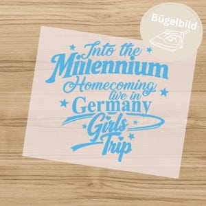 Könnte beinhalten: Ein hellblaues Bügelbild mit dem Text Into the Millennium Homecoming live in Germany Girls Trip, verziert mit Sternen und geschwungenen Linien. Das Design befindet sich auf einer transparenten Folie mit einem runden Aufkleber.