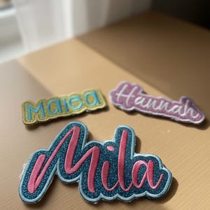 Könnte beinhalten: Drei gestickte Namensaufnäher: "Maica" in Gelb und Blau, "Hannah" in Rosa und "Mila" in Rosa und Türkis. Die Namen sind in Schreibschrift mit Glitzerakzenten, ideal zum Personalisieren von Artikeln.