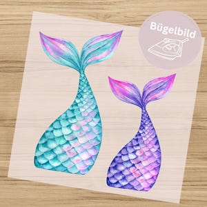 May include: Two watercolour mermaid tail iron-on transfers. One tail is teal and light blue, the other is purple and pink. The tails have a scale pattern. A circular sticker with the text "Bügelbild" and an iron graphic is in the upper right corner.