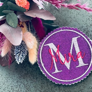 Könnte beinhalten: Runder Filz-Kreis in Lila mit einem weißen gestickten Buchstaben "M" und dem Namen "Mira" in rosa gestickt.