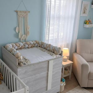 Könnte beinhalten: Ein Wickeltisch in hellem Holzton mit gepolsterter Oberseite und geflochtenem Stoßfänger. Der Wickeltisch hat einen weißen Streifen auf der Vorderseite. Ein weißes Kinderbett ist links zu sehen. Der Raum hat eine hellblaue Wand und einen Makramee-Wandbehang.