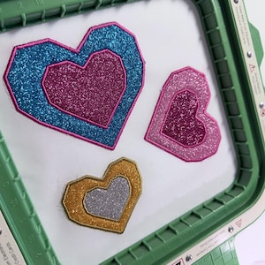 Könnte beinhalten: Drei glitzernde herzförmige Aufnäher in einem grünen Stickrahmen. Das größte Herz ist türkis mit einem rosa Zentrum, das zweite ist rosa und das kleinste ist goldfarben mit einem silbernen Zentrum. Der Rahmen hat den Text "Keep Away From Computer Disks and Hard Drives."