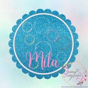 Könnte beinhalten: Ein blaues und weißes Stickdesign mit dem Namen "Mila" in rosa Schrift. Das Design enthält einen Kreis mit einem gewellten Rand und mehreren kleineren Kreisen darin.