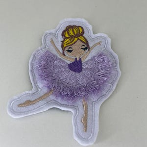 Könnte beinhalten: Gestickter Aufnäher einer Ballerina in einem lila Tutu, die Arme ausgestreckt. Die Ballerina hat blondes Haar und trägt einen lila Body. Der Aufnäher ist weiß umrandet und eignet sich für Kleidung oder Accessoires.