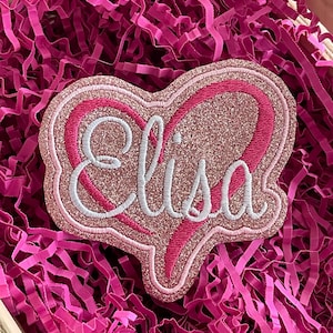 Könnte beinhalten: Ein pinkfarbener, glitzernder herzförmiger Aufnäher mit dem Namen "Elisa" in weißer Schrift gestickt. Der Aufnäher ist von einem pinkfarbenen Rand umgeben und liegt auf einem Bett aus pinkfarbenem Zerkleinerungspapier.