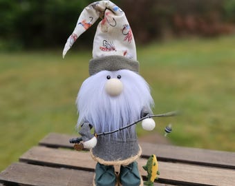May include: A charming gnome figure wearing a grey coat, green boots, and a patterned hat with fishing lures. The gnome has a long white beard, a round nose, and is holding a fishing rod with a bobber and a small yellow fish.