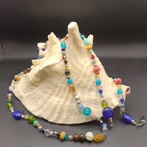 May include: A colorful beaded necklace with various shapes and sizes of beads in shades of blue, green, yellow, red, and clear. The necklace is draped over a large, cream-colored seashell against a dark background.