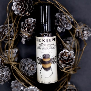 Könnte beinhalten: Eine kleine Glasflasche mit Roll-on-Parfüm, schwarzem Verschluss und weißem Etikett. Das Etikett zeigt eine Comic-Biene mit dem Text "Bee Keeper Roll-on Perfume Honey Amber Orange" und "By Scream Printables". Die Flasche ist von einem Kranz aus Tannenzapfen umgeben.