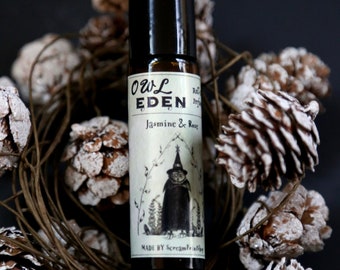 May include: A small brown glass bottle with a black cap and a white label. The label says "Owl Eden Roll-on Perfume Jasmine & Rose" and features a black and white illustration of an owl wearing a witch's hat. The label also says "Made by ScreamFruitShop."