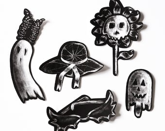 May include: Black and white illustrations of Halloween-themed objects. Includes a ghost wearing a crown, a sun hat with a ribbon, a skull flower, a pair of bat wings, and a ghost with a jack-o'-lantern face.