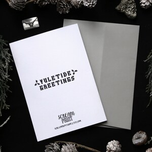 May include: A white card with black text that reads "Yuletide Greetings" and "Scream Print" with the website address "screamprint.etsy.com". The card is on a black background with a gray envelope.