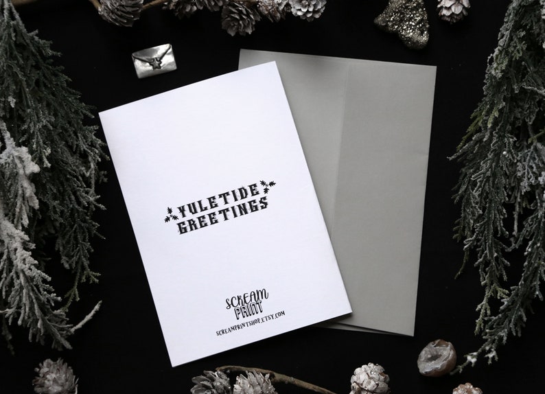 May include: A white card with black text that reads "Yuletide Greetings" and "Scream Print" with the website address "screamprint.etsy.com". The card is on a black background with a gray envelope.