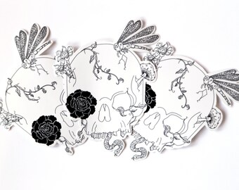 Op de afbeelding: Drie zwart-witte schedel-illustraties met bloem- en libellen-details. Elke schedel is omgeven door ranken, bloemen en libellen.