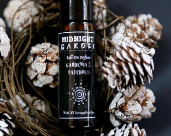 May include: A black roll-on perfume bottle with a white label that reads "Midnight Garden Roll-On Perfume Gardenia & Patchouli Made by ScreamPrinting". The bottle is surrounded by white pine cones.