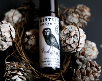 May include: A glass roller bottle with a black cap and a white label. The label features a black and white illustration of an owl with the text "Winter Solstice" and a list of ingredients: "Pine, Fresh Soil, Frankincense, Myrrh". The bottle is surrounded by pine cones.
