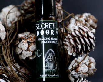 May include: A small black roll-on bottle with a green label that says "SECRET DOOR" and "DRAGONS BLOOD & POMEGRANATE". The label also features a small illustration of a door with a crescent moon above it. The bottle is surrounded by white pine cones.