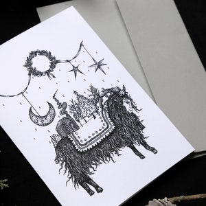 May include: A black and white illustration of a goat with a wreath, stars, a crescent moon, and a small house hanging from a string. The goat is standing on a snowy ground with a pine tree in the background.