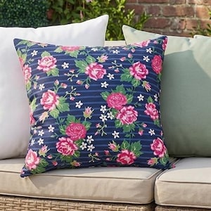 May include: A square throw pillow featuring a dark blue background with thin horizontal white stripes and a repeating pattern of pink roses and small white flowers. The pillow is positioned on a beige wicker outdoor sofa with cushions.