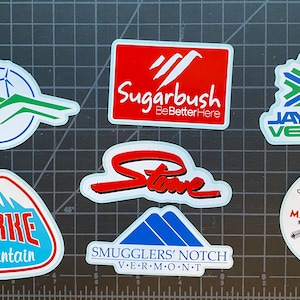 May include: A collection of seven colorful ski resort stickers. The stickers feature the names of the resorts, including Burke Mountain, Sugarbush, Jay Peak Vermont, Stowe, Smugglers' Notch Vermont, and Mad River Glen.