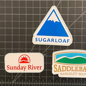 May include: Three white stickers with mountain logos. The top sticker is blue with a white mountain peak and the text "SUGARLOAF". The left sticker has a red sun rising over a mountain and the text "Sunday River". The right sticker has a green mountain with a blue sky and the text "SADDLEBACK RANGELEY - MAINE".