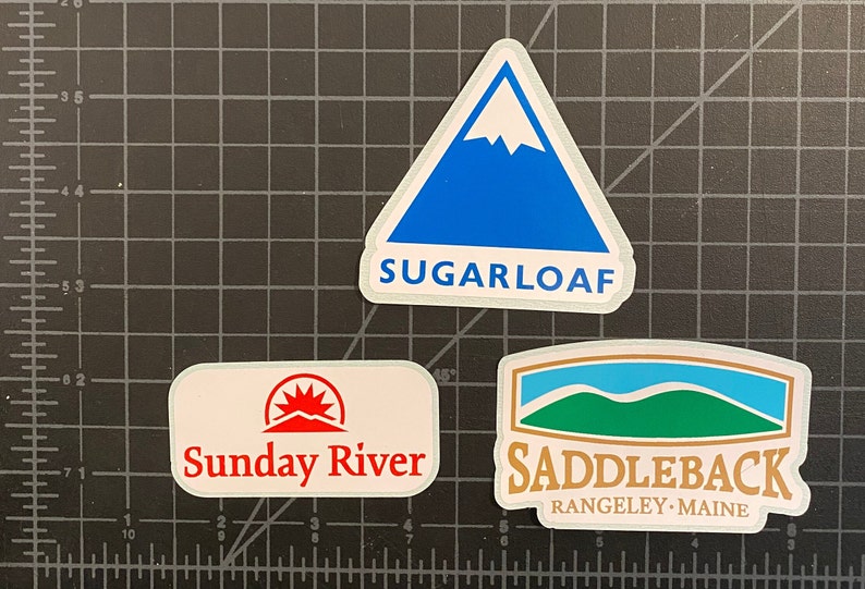May include: Three white stickers with mountain logos. The top sticker is blue with a white mountain peak and the text "SUGARLOAF". The left sticker has a red sun rising over a mountain and the text "Sunday River". The right sticker has a green mountain with a blue sky and the text "SADDLEBACK RANGELEY - MAINE".