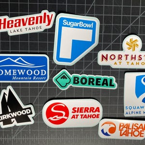 May include: A collection of ten colorful stickers featuring the names of different ski resorts in Lake Tahoe, California. The stickers are white with black, red, blue, and green lettering. The stickers include the names: Heavenly, SugarBowl, Homewood, Boreal, Northstar, Kirkwood, Sierra at Tahoe, Palisades Tahoe, and Squaw Valley Alpine Meadows.