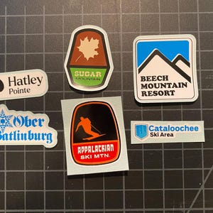 May include: A collection of six colorful ski resort stickers. The stickers feature the names of the resorts, including Sugar Mountain, Beech Mountain Resort, Ober Gatlinburg, Hatley Pointe, Appalachian Ski Mtn., and Cataloochee Ski Area.