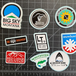 May include: A collection of nine colorful stickers featuring the names of different ski resorts in Montana. The stickers are all round or oval shaped and have a white background with black text. The stickers are arranged in a grid pattern on a gray surface.