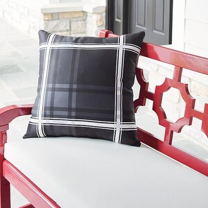 May include: A square outdoor pillow with a black and gray plaid pattern, accented with white stripes. The pillow is placed on a white cushion of a red bench. The bench has a decorative geometric design.