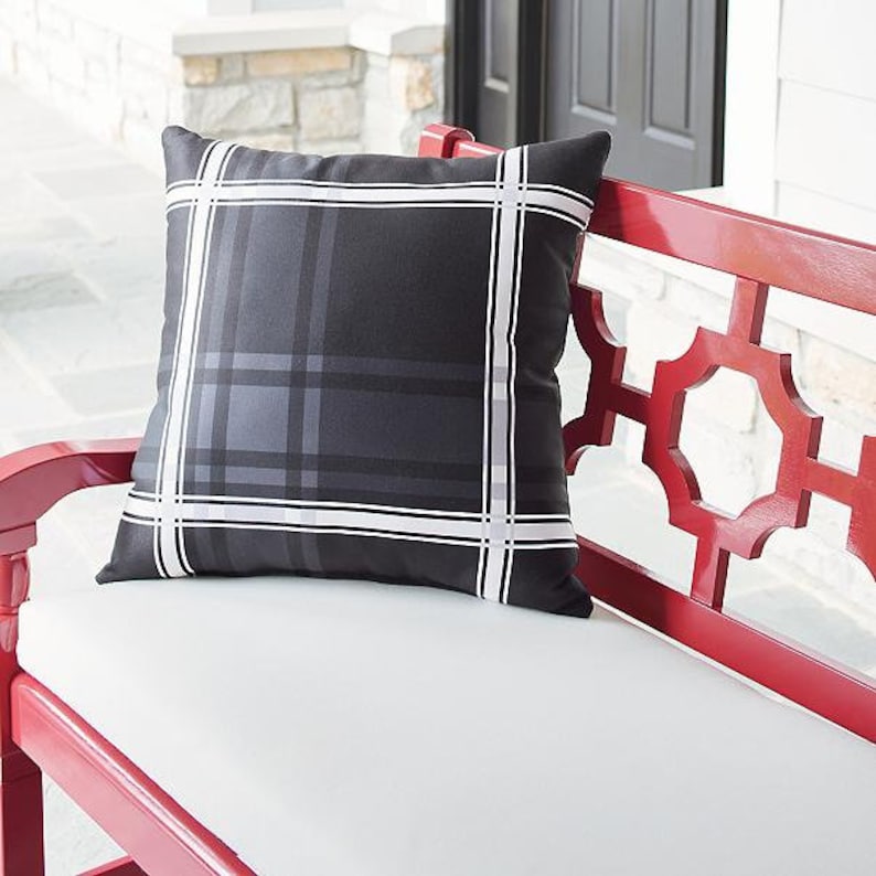 May include: A square outdoor pillow with a black and gray plaid pattern, accented with white stripes. The pillow is placed on a white cushion of a red bench. The bench has a decorative geometric design.