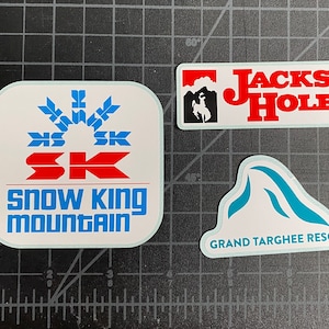 May include: Three white stickers with ski resort logos. One features "Snow King Mountain" in blue and red. Another says "Jackson Hole" with a cowboy silhouette. The third reads "Grand Targhee Resort" with a mountain graphic.