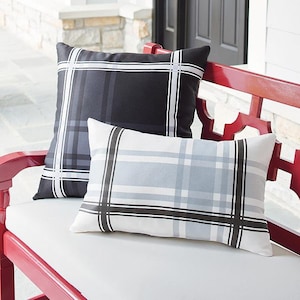 May include: Two decorative outdoor pillows with a plaid pattern. One pillow is black with gray and white stripes, and the other is white with gray and black stripes. The pillows are rectangular and sit on a red bench.
