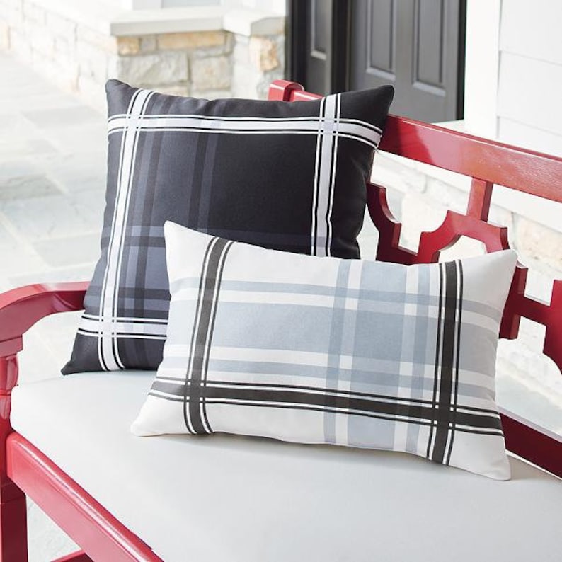 May include: Two decorative outdoor pillows with a plaid pattern. One pillow is black with gray and white stripes, and the other is white with gray and black stripes. The pillows are rectangular and sit on a red bench.