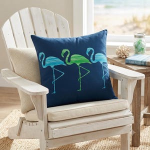 May include: A square navy blue throw pillow featuring a graphic print of three flamingos standing on one leg. The flamingos are rendered in shades of teal, turquoise, and lime green with a horizontal striped texture throughout their silhouettes.
