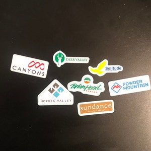 May include: A collection of colorful stickers featuring the names of various ski resorts, including Canyon, Deer Valley, Brian Head, Nordic Valley, Solitude, Sundance, and Powder Mountain.