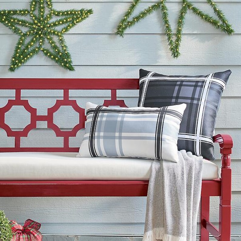 May include: A red bench with white cushions and two plaid pillows in shades of gray and blue. A gray throw blanket with fringe is draped over the bench. Decorative star-shaped lights and greenery are in the background.