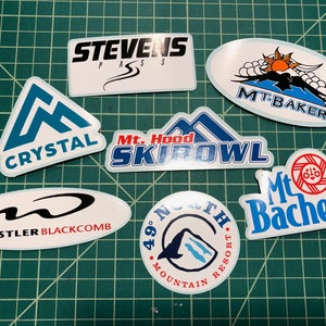 May include: A collection of nine colorful ski resort stickers. The stickers feature the names of various ski resorts, including Stevens Pass, Crystal, Mt. Hood Ski Bowl, Mt. Baker, Whistler Blackcomb, 49° North Mountain Resort, and Mt. Bachelor.